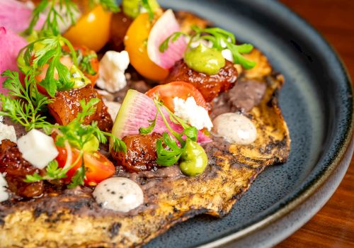 A savory flatbread topped with colorful veggies, herbs, and dollops of sauce on a dark plate. _end of sentence_