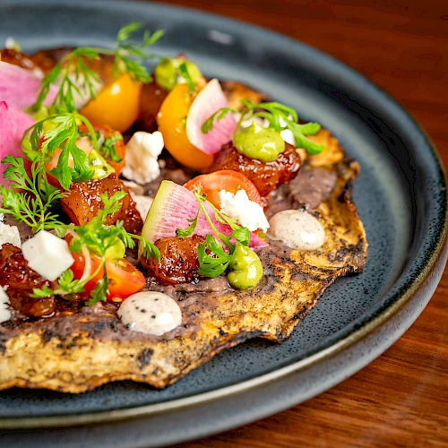 A savory flatbread topped with colorful veggies, herbs, and dollops of sauce on a dark plate. _end of sentence_