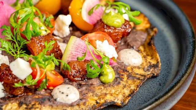 A savory flatbread topped with colorful veggies, herbs, and dollops of sauce on a dark plate. _end of sentence_