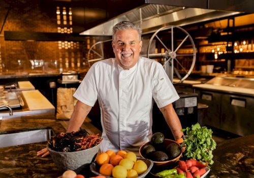 A chef in a white coat stands behind a kitchen counter with fresh fruit, vegetables, and herbs, smiling in a busy restaurant kitchen.