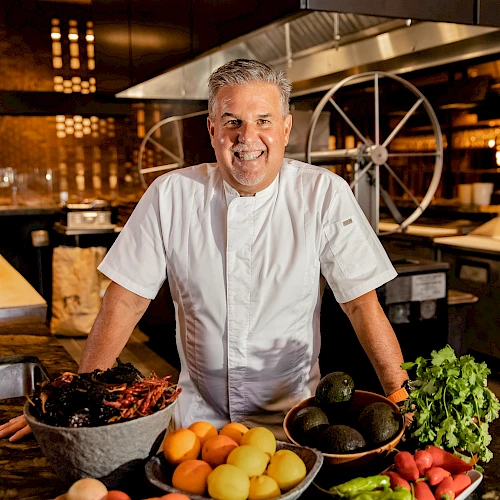 A chef in a white coat stands behind a kitchen counter with fresh fruit, vegetables, and herbs, smiling in a busy restaurant kitchen.