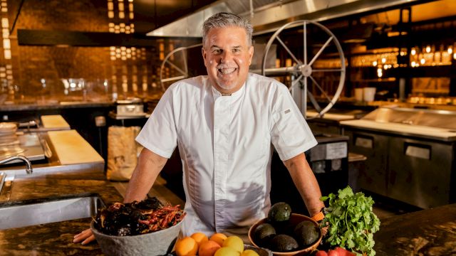 A chef in a white coat stands behind a kitchen counter with fresh fruit, vegetables, and herbs, smiling in a busy restaurant kitchen.