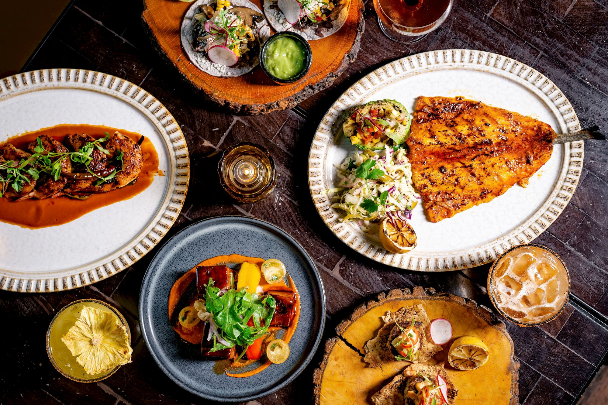 A spread of plated dishes: tacos, grilled chicken, vibrant veggies, sauces, and drinks on a dark table, ready-to-eat feast.
