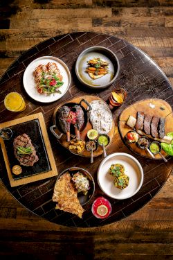A round table with assorted dishes: steaks, slices of bread, small plates of sides, sauces, and a few greens, all on a dark wooden floor, beautifully plated.