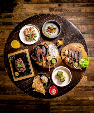 A round table with assorted dishes: steaks, slices of bread, small plates of sides, sauces, and a few greens, all on a dark wooden floor, beautifully plated.