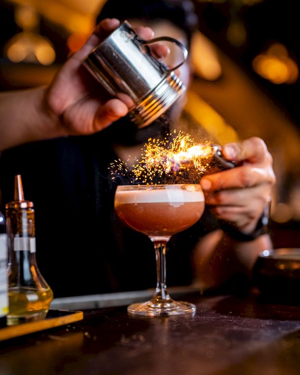 A bartender sprinkles a sparkler on a frothy cocktail, pouring with a shaker in a dim, warm-lit bar.