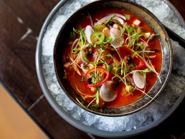 A vibrant seafood soup or bouillabaisse served in a round bowl on ice; bright red broth, scallions, herbs, and small fish or shellfish pieces. End.