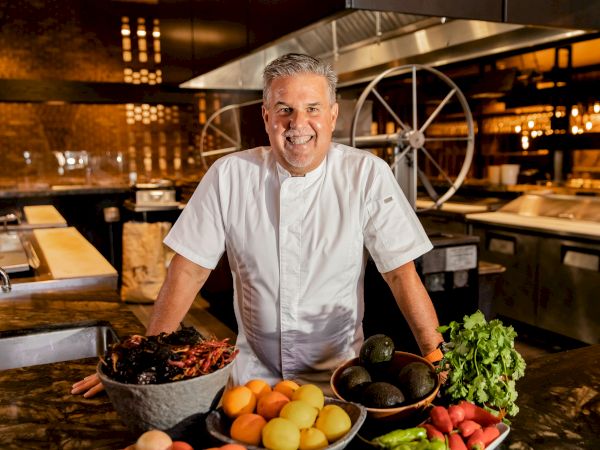 A chef in a white coat stands behind a counter with fresh produce&mdash;peppers, peaches, avocados, greens, and other colorful ingredients. The kitchen glows warmly.