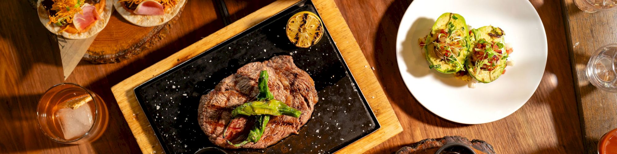 A variety of plated dishes on a wooden table: tacos, a sizzling steak on a hot board, tacos, bowls, a pizza or flatbread, salads, and drinks.