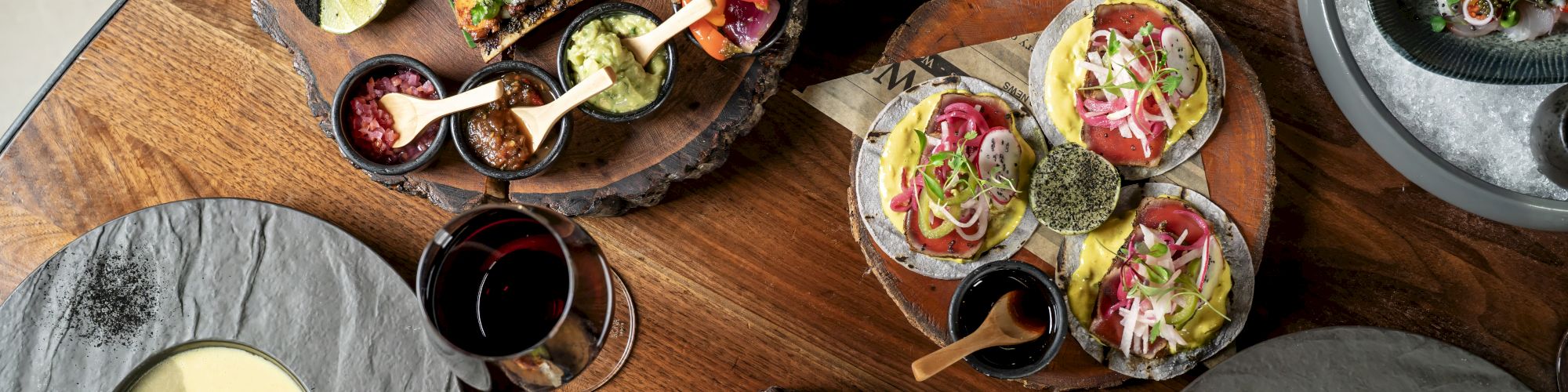 A spread of tapas and small plates: sliders, tacos, deviled eggs, greens, a taco cone, bowls, sauces, and wine on a wooden table, tasty bites everywhere.