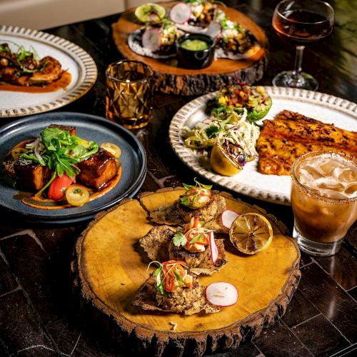 A spread of gourmet dishes on wooden platters and plates, with drinks, greens, and vibrant toppings, all artfully arranged for a stylish feast.