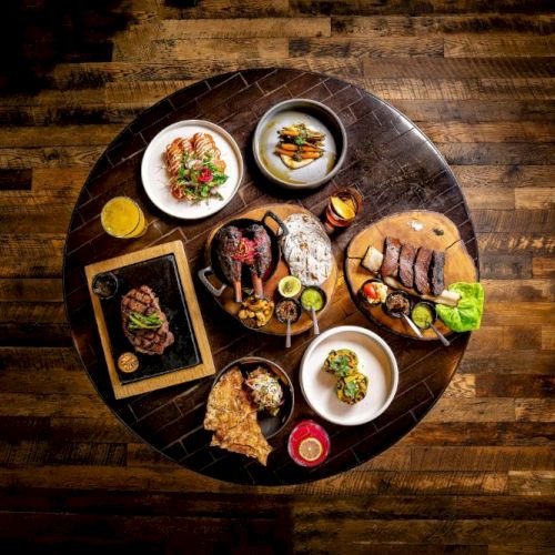 A round table with wood flooring shows a feast: steaks, burgers, ribs, salads, sauces, grilled veggies, and small plates around a central spread, delicious.