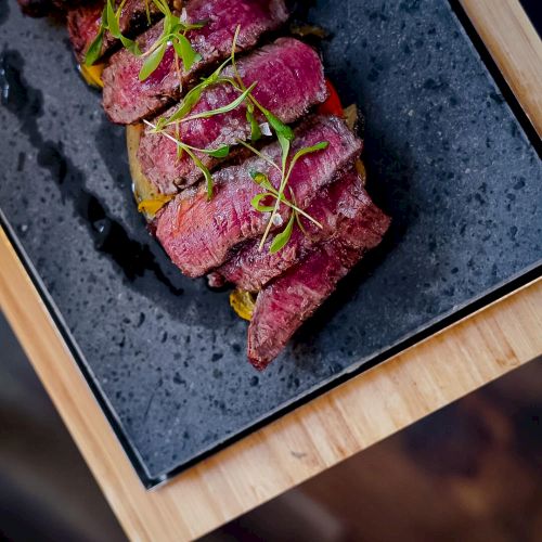 Sliced seared beef tataki on a dark plate, garnished with microgreens, resting on a wooden board with chopsticks&mdash;salty, smoky, fresh.