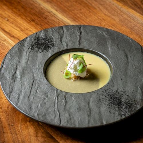 A black slate plate with a small, elegant appetizer in the center, featuring a pale sauce pool and a tiny green-turred garnish on top.