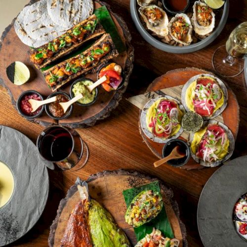 A spread of fancy tapas and small plates: tacos, deviled eggs, skewers, ceviche bowls, and colorful desserts on wooden boards and slate plates.