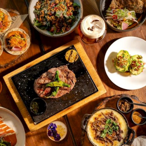 A variety of plated dishes and a sizzling steak on a carved tabletop, with bowls and sides around a shared feast, delicious and colorful.