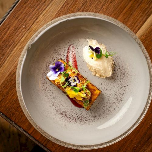 A plated dessert: a small portion with edible flowers and sauce on a bread-like base beside a scoop of pale cream, all artfully arranged on a round plate.