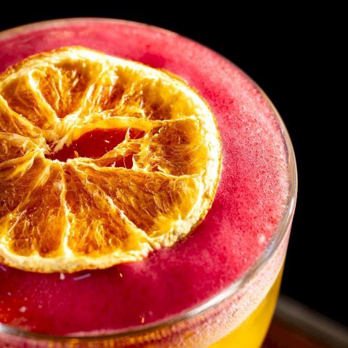 A vibrant smoothie or juice with a bright yellow-orange base and a magenta-pink foam top, garnished with a dried orange slice on the rim.