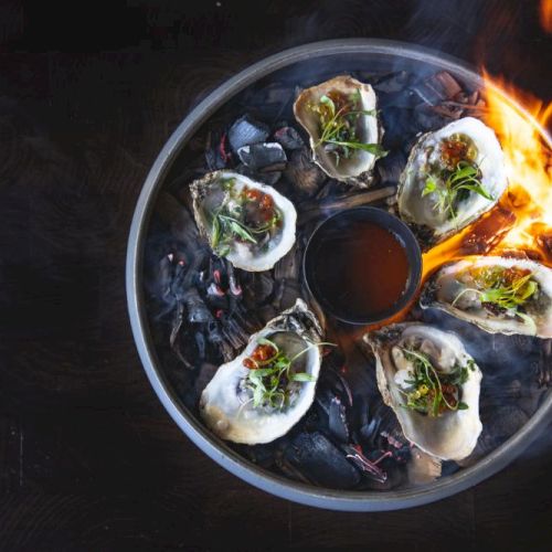 A circular pan with six neatly arranged dumplings or seafood bites, garnished, placed on a dark surface, with flames licking the side of the pan, creating a dramatic, fiery scene.