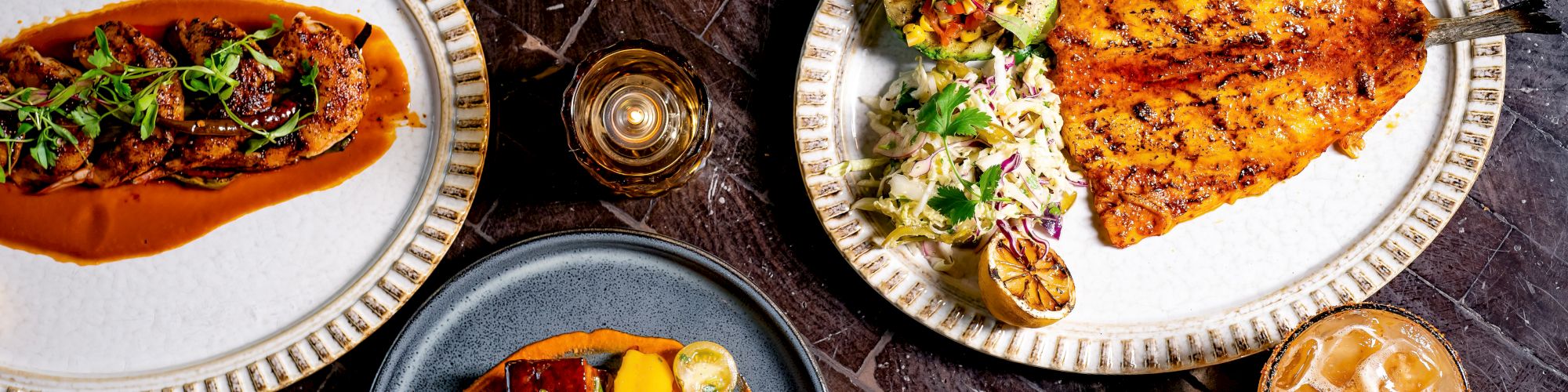 A top-down view of a table full of plated dishes: tacos, roast meat with sauce, a ceviche-like plate, burgers, plus sauces and drinks, all arranged appetizingly.