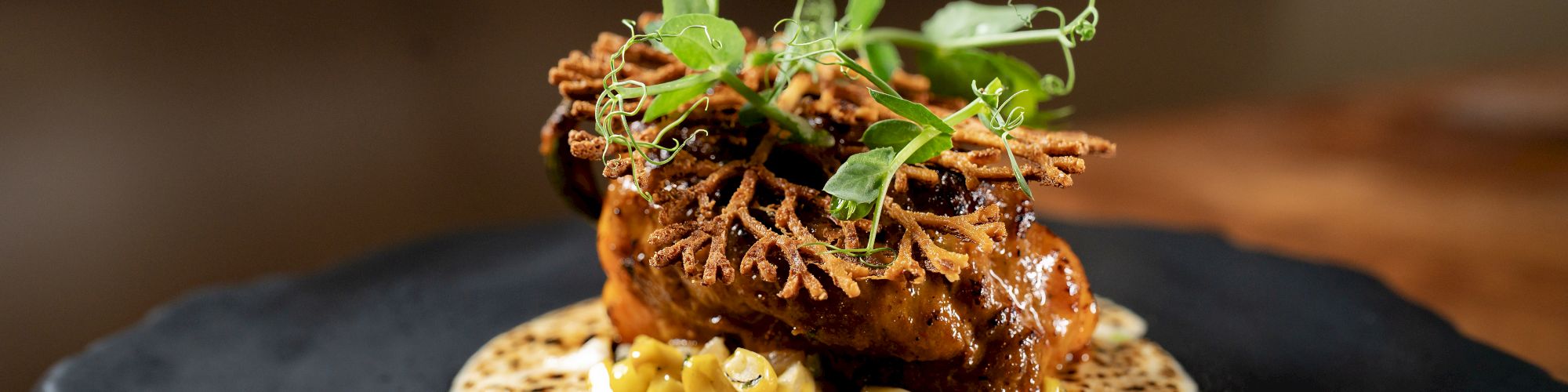 A plated dish with a crispy tortilla base, topped by a seared meat patty, crunchy greens, and a glossy sauce on a dark plate.